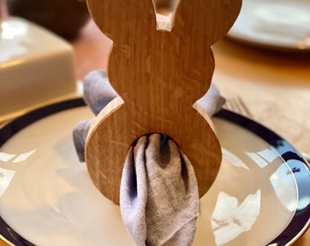 Puede incluir: Anillo de servilleta de madera en forma de conejo con una servilleta gris en su interior. El anillo está sobre un plato blanco con un borde azul.