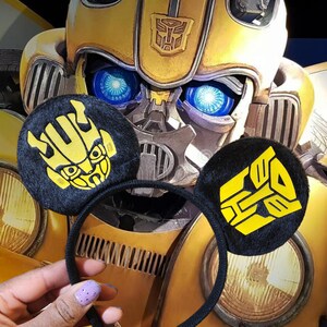 May include: Black headband with yellow fuzzy ears featuring the Transformers logo. The ears are shaped like Mickey Mouse ears. The headband is in front of a large yellow and black robot.