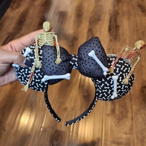 May include: Black and white patterned Minnie Mouse ears with a black headband. Two skeleton figures are attached to the ears, one on each side. The skeletons are made of plastic and have white bones.