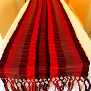 May include: A long, rectangular table runner with vertical stripes in shades of red, brown, and black. The runner has a fringed edge and is displayed on a white surface. The textile is likely woven.