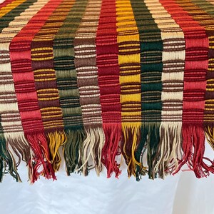 May include: A vibrant woven table runner featuring a striped design in red, green, yellow, and brown. The runner has a fringed edge and is likely made of fabric. The pattern combines horizontal and vertical stripes, creating a textured look.