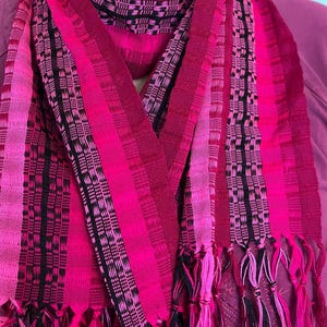May include: A vibrant pink scarf with a striped pattern and fringe. The scarf features varying shades of pink and black woven stripes. The fringe at the ends adds a decorative touch to this textile accessory.