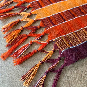 May include: A woven textile runner featuring alternating vertical stripes in shades of deep burgundy, bright orange, and golden yellow. The fabric is finished with long, hand-knotted fringe tassels at the edge, incorporating the same color palette.