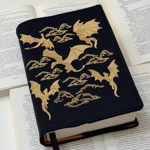 Könnte beinhalten: Schwarzer Buchumschlag mit goldgestickten Drachen und Wolken. Der Umschlag liegt auf einem offenen Buch mit weißen Seiten. Ein schwarzes Lesezeichenband ist sichtbar.