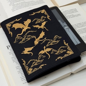 Op de afbeelding: Een zwarte Kindle-hoes met goud geborduurde draken en wolken. De hoes is rechthoekig en heeft een gedetailleerd ontwerp. De hoes ligt op een open boek met witte pagina's.