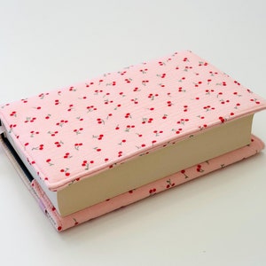Könnte beinhalten: Ein rosa Buchcover mit einem Kirschendruck. Der Bezug ist aus Stoff und hat ein weißes Band als Lesezeichen.