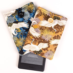 以下が含まれることがあります： 日本の鶴と花の模様が施された、布製の電子書籍リーダーカバー2つ。1つは青と黒、もう1つは茶色と金色。両方のカバーにはボタン留めがあります。