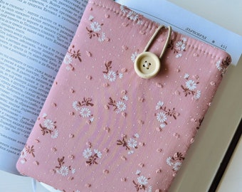 May include: A pink book cover with a white floral pattern and a white button closure. The cover has a white elastic strap.