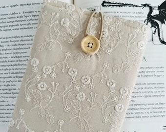Puede incluir: Una bolsa de cordón beige con un diseño de bordado floral blanco. La bolsa tiene un cierre de botón de madera y está encima de un libro.
