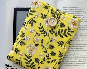 May include: A yellow floral fabric e-reader cover with a white button closure. The cover has a floral pattern with white flowers and green leaves.