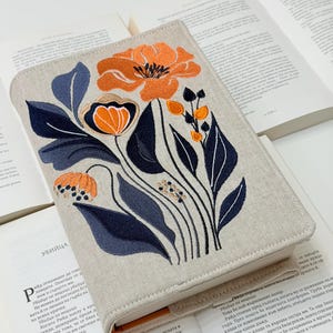 Könnte beinhalten: Beige Buchhülle mit einem gestickten Blumendesign mit orangefarbenen und marineblauen Blumen und Blättern. Die Hülle liegt auf offenen Büchern.