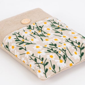 K&ouml;nnte beinhalten: Eine beige Stoffh&uuml;lle mit einem floralen G&auml;nsebl&uuml;mchen-Stickdesign. Die H&uuml;lle hat einen Knopfverschluss und eine rechteckige Form. Die gestickten G&auml;nsebl&uuml;mchen haben wei&szlig;e Bl&uuml;tenbl&auml;tter und gelbe Zentren mit gr&uuml;nen Stielen und Bl&auml;ttern.