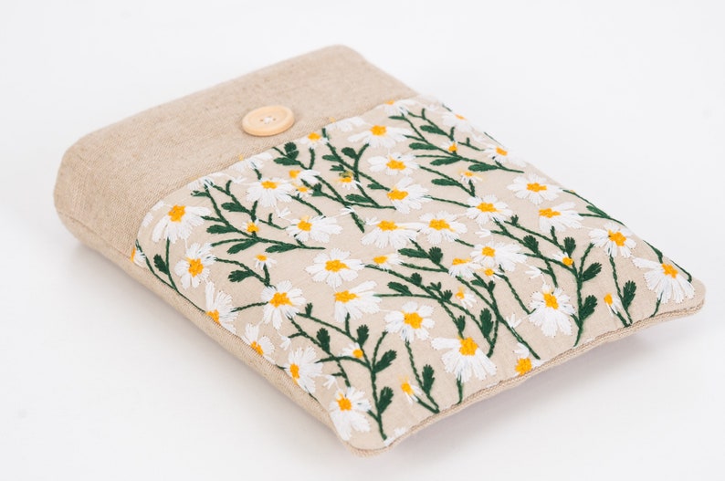 K&ouml;nnte beinhalten: Eine beige Stoffh&uuml;lle mit einem floralen G&auml;nsebl&uuml;mchen-Stickdesign. Die H&uuml;lle hat einen Knopfverschluss und eine rechteckige Form. Die gestickten G&auml;nsebl&uuml;mchen haben wei&szlig;e Bl&uuml;tenbl&auml;tter und gelbe Zentren mit gr&uuml;nen Stielen und Bl&auml;ttern.