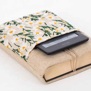 K&ouml;nnte beinhalten: Ein beiger Buchumschlag mit einem floralen G&auml;nsebl&uuml;mchenmuster auf der oberen Klappe, der einen Kindle E-Reader h&auml;lt. Der Umschlag besteht aus einem nat&uuml;rlich aussehenden Stoff und ist mit einer Schnur gebunden.