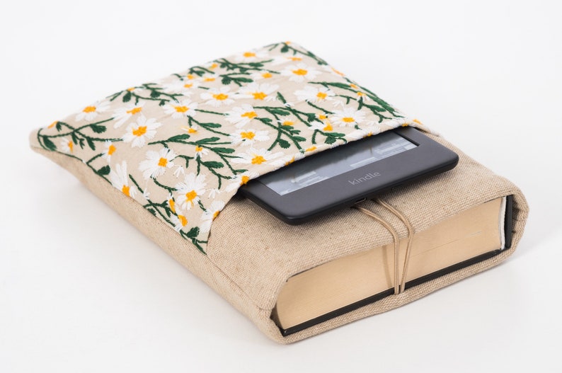 K&ouml;nnte beinhalten: Ein beiger Buchumschlag mit einem floralen G&auml;nsebl&uuml;mchenmuster auf der oberen Klappe, der einen Kindle E-Reader h&auml;lt. Der Umschlag besteht aus einem nat&uuml;rlich aussehenden Stoff und ist mit einer Schnur gebunden.