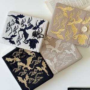 May include: Four square book covers in off-white, tan, and black. Each cover features an embroidered design of bats and clouds. The off-white cover has navy blue embroidery, the tan covers have gold and white embroidery, and the black cover has gold embroidery.