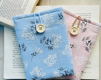 May include: Two fabric phone cases with floral patterns. The case on the left is light blue with white flowers and a black button. The case on the right is pink with white flowers and a white button.