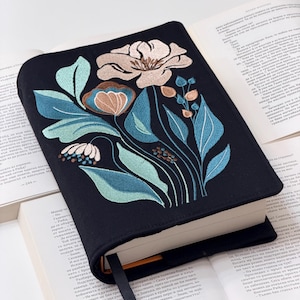 May include: A black book cover embroidered with a floral design in blue, beige, and white. The embroidery depicts flowers and foliage. A black ribbon bookmark is present. The book is open, displaying text.
