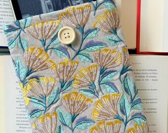 May include: Beige tablet sleeve with embroidered yellow and teal floral design. Features a wooden button closure. The sleeve is open, revealing a tablet screen. The background includes open books.