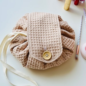 May include: A beige waffle-textured drawstring makeup bag with a single button closure and a white cotton strap.