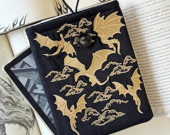 Op de afbeelding: Zwarte e-reader hoes met gouden geborduurde draken en wolken. De hoes heeft een zwarte knoopsluiting en een zwarte koordlus. De e-reader is gedeeltelijk zichtbaar in de hoes, rustend op open boeken.