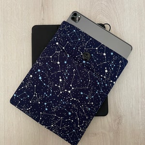 以下が含まれることがあります： 白い星座柄のネイビーブルーのタブレットスリーブで、ボタン留めになっています。スリーブはタブレットまたはiPadに合うように設計されています。