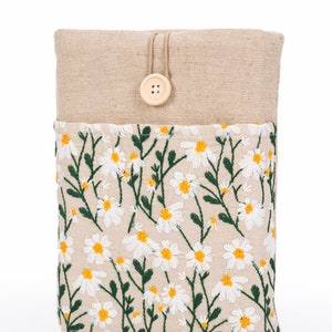 K&ouml;nnte beinhalten: Eine beige Stoffh&uuml;lle mit einem gestickten G&auml;nsebl&uuml;mchen-Blumenmuster. Die H&uuml;lle hat einen Knopfverschluss und besteht aus einem naturfarbenen, leinenartigen Material. Das G&auml;nsebl&uuml;mchenmuster hat wei&szlig;e Bl&uuml;tenbl&auml;tter und gelbe Zentren mit gr&uuml;nen Stielen und Bl&auml;ttern.
