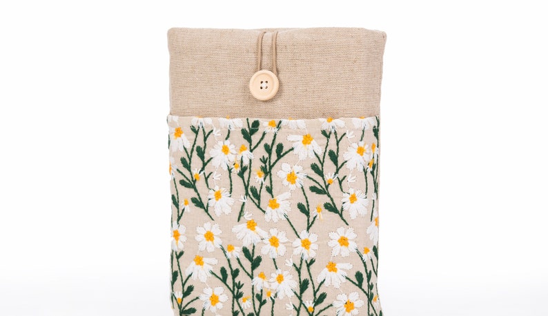 K&ouml;nnte beinhalten: Eine beige Stoffh&uuml;lle mit einem gestickten G&auml;nsebl&uuml;mchen-Blumenmuster. Die H&uuml;lle hat einen Knopfverschluss und besteht aus einem naturfarbenen, leinenartigen Material. Das G&auml;nsebl&uuml;mchenmuster hat wei&szlig;e Bl&uuml;tenbl&auml;tter und gelbe Zentren mit gr&uuml;nen Stielen und Bl&auml;ttern.
