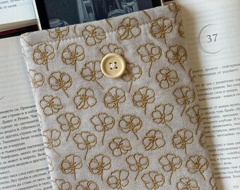 May include: Beige fabric e-reader sleeve with a repeating gold floral embroidery pattern. The sleeve has a button closure and is resting on an open book.