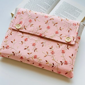 Könnte beinhalten: Eine rosa Buchhülle mit floraler Stickerei. Der Bezug weist ein sich wiederholendes Muster aus kleinen roten Blüten und braunen Blättern auf. Zwei große, runde, beige Knöpfe sichern die Klappe. Die Hülle ist offen und zeigt die Seiten eines Buches.