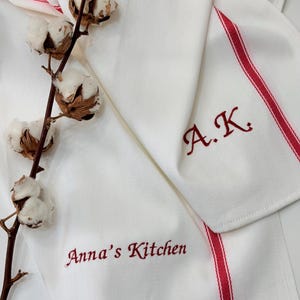 Può includere: Strofinacci da cucina bianchi con strisce rosse e testo ricamato. Uno strofinaccio recita "Anna's Kitchen" e l'altro ha le iniziali "A.K.". Un rametto di cotone aggiunge un tocco rustico alla scena.