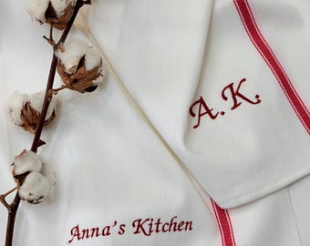 May include: White kitchen towels with red stripes and embroidered text. One towel reads "Anna's Kitchen" and the other has the initials "A.K." A sprig of cotton adds a rustic touch to the scene.