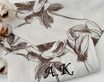 May include: Off-white linen table runner and matching napkin set. The set features a brown floral sketch design. The napkin is embroidered with the initials "A.K." in black. Cotton bolls are visible in the background.