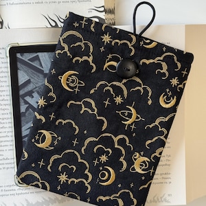 May include: Black e-reader case with a celestial pattern. Gold embroidered clouds, moons, stars, and crosses decorate the fabric. A black button and elastic loop fasten the cover. The e-reader is partially visible.