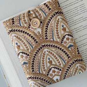 Può includere: Una copertina per libro bianca con chiusura a bottone marrone. La copertina è decorata con un motivo ripetitivo di archi ricamati in tonalità di marrone, oro e blu.