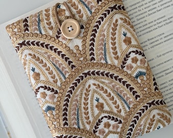May include: A white book cover with a brown button closure. The cover is decorated with a repeating pattern of embroidered arches in shades of brown, gold, and blue.