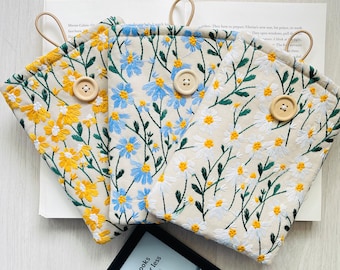 May include: Three different floral patterned fabric pouches with a button closure. The pouches are beige with yellow, blue, and white flowers. The pouches are laying on a white surface with a black e-reader on top.