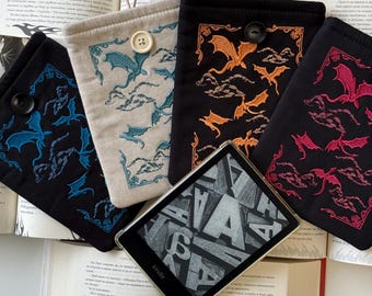 Op de afbeelding: Vier verschillende e-reader hoesjes met geborduurde drakenontwerpen. De hoesjes zijn in zwart, beige en rood. De hoesjes hebben een knoopsluiting. De hoesjes liggen op een oppervlak met een boek en een e-reader.