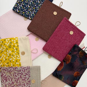 Può includere: Una collezione di copertine per libri in tessuto di vari colori e motivi. I disegni includono trame floreali, a nido d'ape e a spina di pesce. Ogni copertina presenta una chiusura con bottone in legno e un anello.