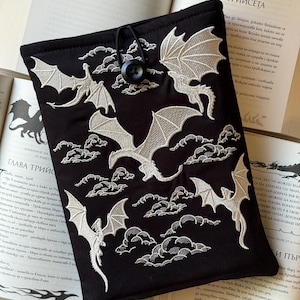 Può includere: Custodia per libri nera con draghi e nuvole bianche ricamati. La custodia ha una chiusura a bottone ed è appoggiata su libri aperti. Un accessorio a tema fantasy per gli amanti dei libri.