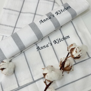 May include: White kitchen towels with grey stripes and a grid pattern. The text "Anna's Kitchen" is embroidered on the rolled towel. A sprig of cotton adds a natural element to the scene.