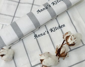 May include: White kitchen towels with gray stripes and a grid pattern. The text "Anna's Kitchen" is embroidered on the rolled towel. A sprig of cotton adds a natural element to the scene.