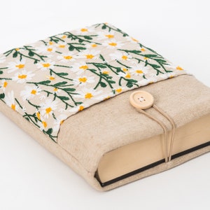 K&ouml;nnte beinhalten: Ein beiger Buchumschlag mit einem gestickten Blumenmuster aus wei&szlig;en G&auml;nsebl&uuml;mchen mit gelben Zentren und gr&uuml;nen Bl&auml;ttern. Der Umschlag hat einen Knopfverschluss und ist aus Leinenstoff.