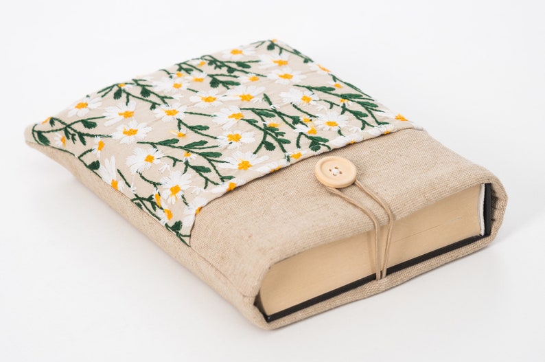 K&ouml;nnte beinhalten: Ein beiger Buchumschlag mit einem gestickten Blumenmuster aus wei&szlig;en G&auml;nsebl&uuml;mchen mit gelben Zentren und gr&uuml;nen Bl&auml;ttern. Der Umschlag hat einen Knopfverschluss und ist aus Leinenstoff.