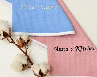 May include: Two waffle-weave kitchen towels, one light blue and one pink, with white trim. The towels are embroidered with "Anna's Kitchen" in a script font. A sprig of cotton adds a natural element to the composition.