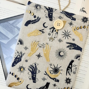 Op de afbeelding: Beige stoffen boekenhoes met een hemels patroon van handen, manen, zonnen en sterren in marineblauwe en gouden borduursels. Voorzien van een knoopsluiting en een lus om te dragen. Ideaal voor het beschermen van boeken of e-readers.