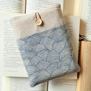 Könnte beinhalten: Eine beige Leinen-Tasche mit einem blauen und weißen Wellenmuster. Die Tasche hat einen braunen Lederriemen und einen Knopf aus Holz.