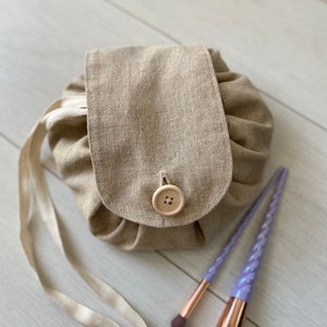 Op de afbeelding: Een beige linnen toilettas met een houten knoopsluiting en een lange beige lintband. Twee paarse make-upkwasten met regenboogharen liggen naast de tas.