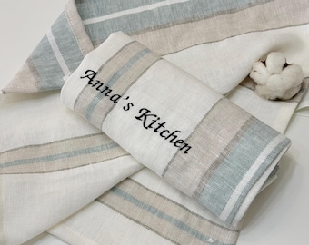 May include: White kitchen towel with blue and tan stripes. The towel is folded and rolled, with the embroidered text "Anna's Kitchen" in black. A cotton boll is in the background. The towel is made of a soft, woven material.