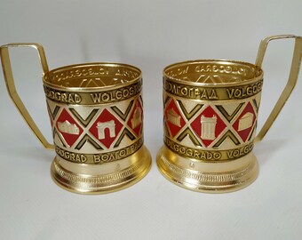 May include: Two gold-colored metal cup holders with a red and gold design. The holders feature the word "Volgograd" written around the top and bottom of the holder. The holders have a handle and a round base.